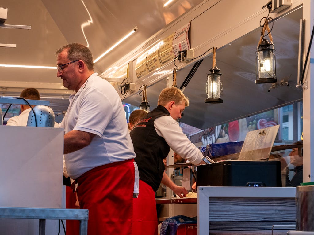 Street food vendor in red apron prepares meals inside a food truck. Busy evening scene.