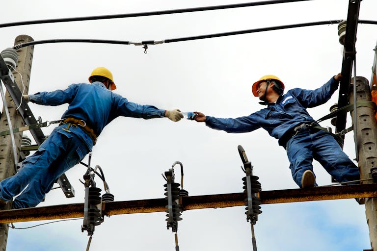 Two electrical workers in blue uniforms collaborating on power lines during daytime.