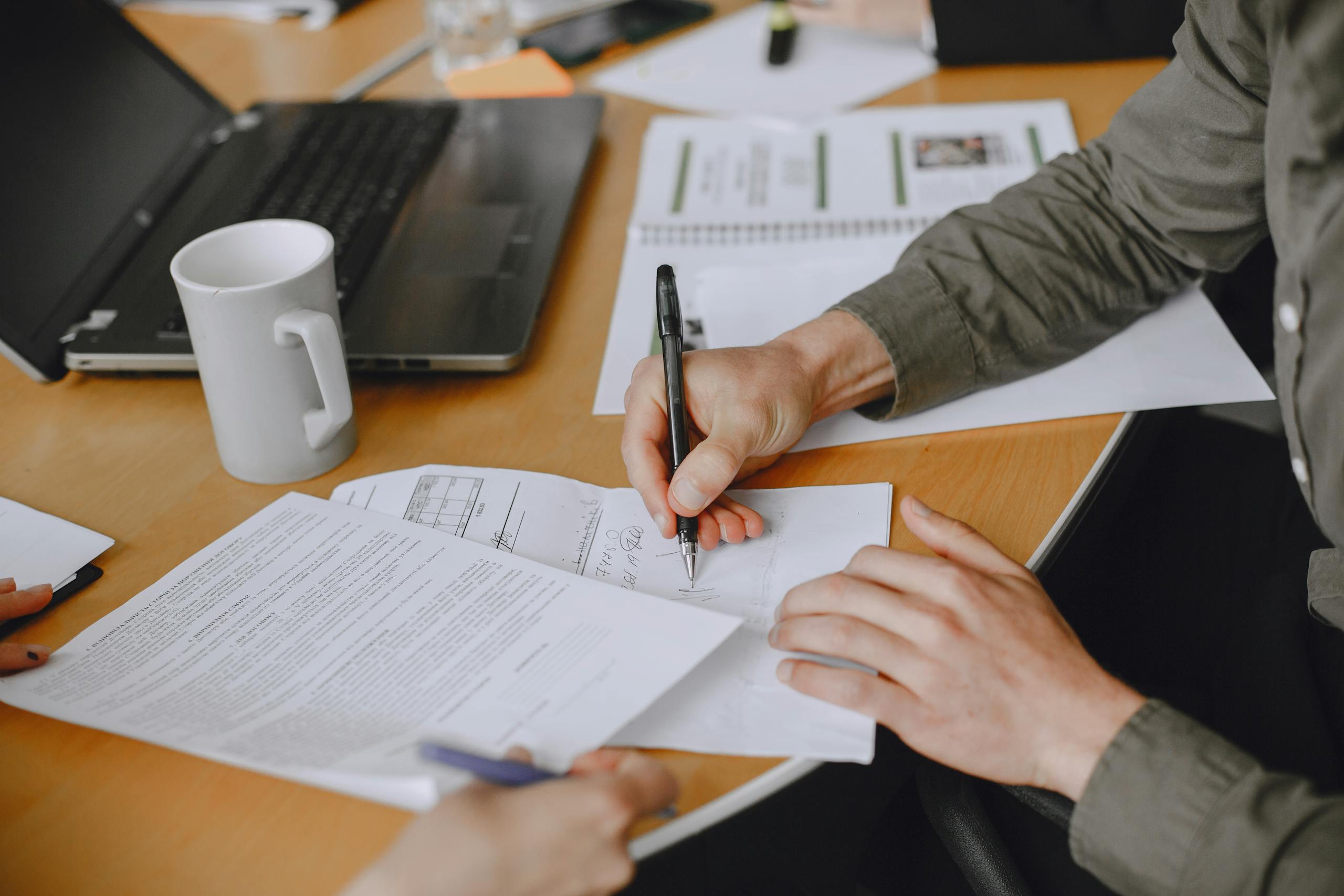 Close-up of individuals signing documents at a business meeting with a laptop on a table.