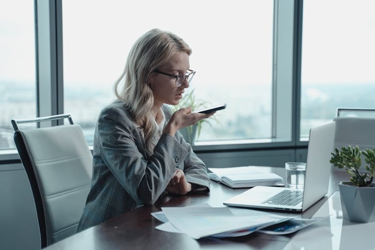 Young businesswoman with blonde hair using a smartphone for speech-to-text typing in a modern office setting.