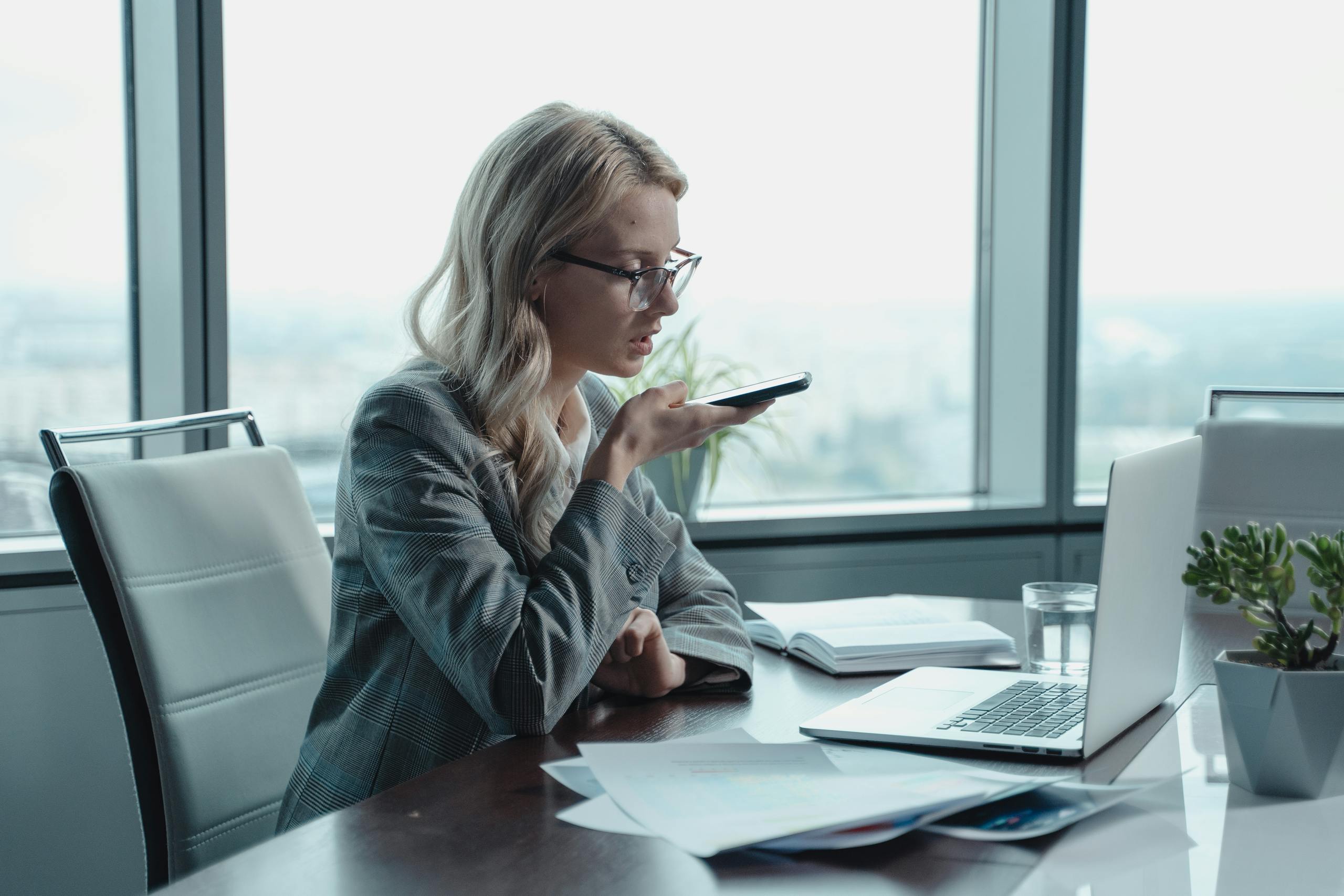 Young businesswoman with blonde hair using a smartphone for speech-to-text typing in a modern office setting.