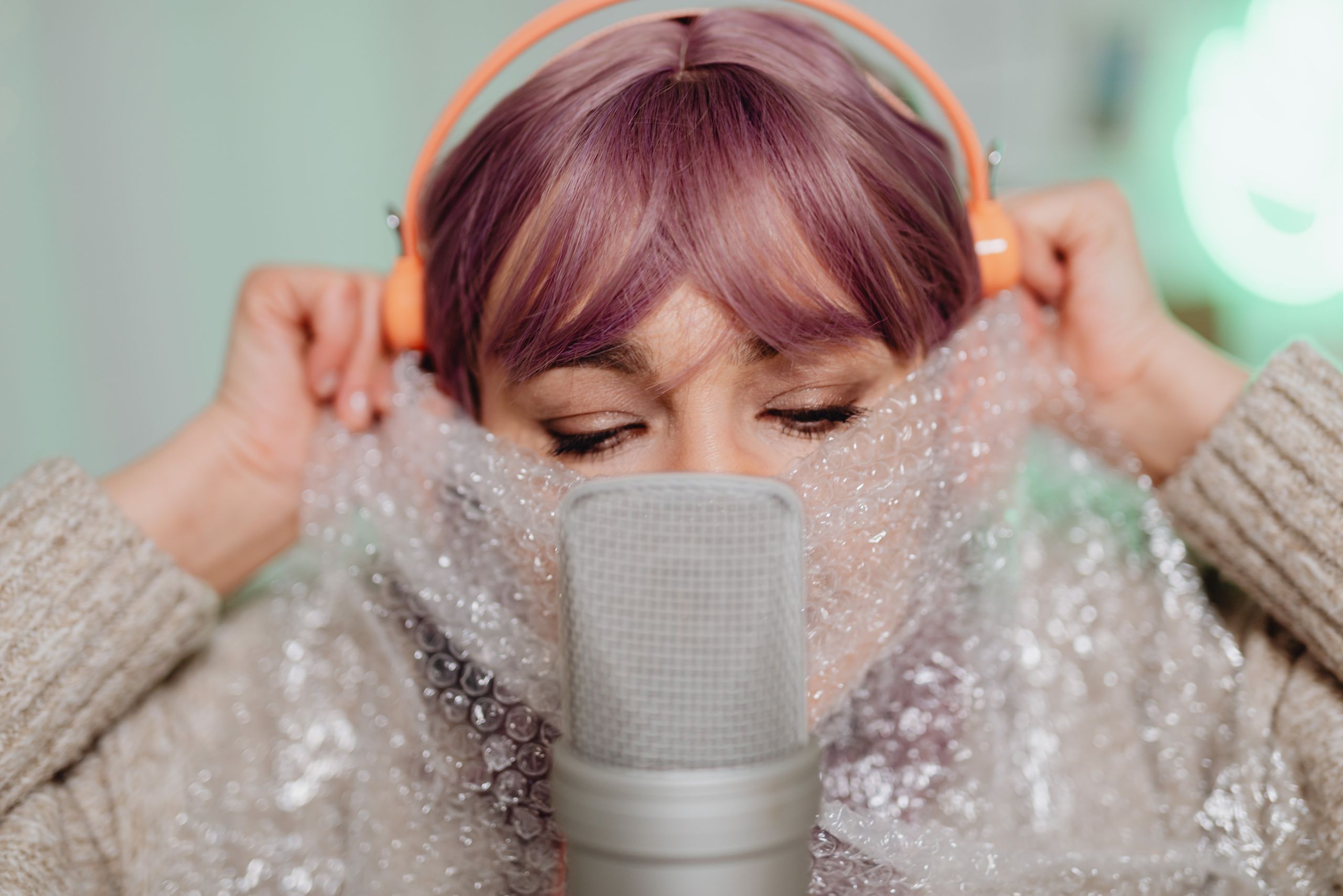 A woman with colored hair and headphones artistically poses with bubble wrap in front of a microphone.