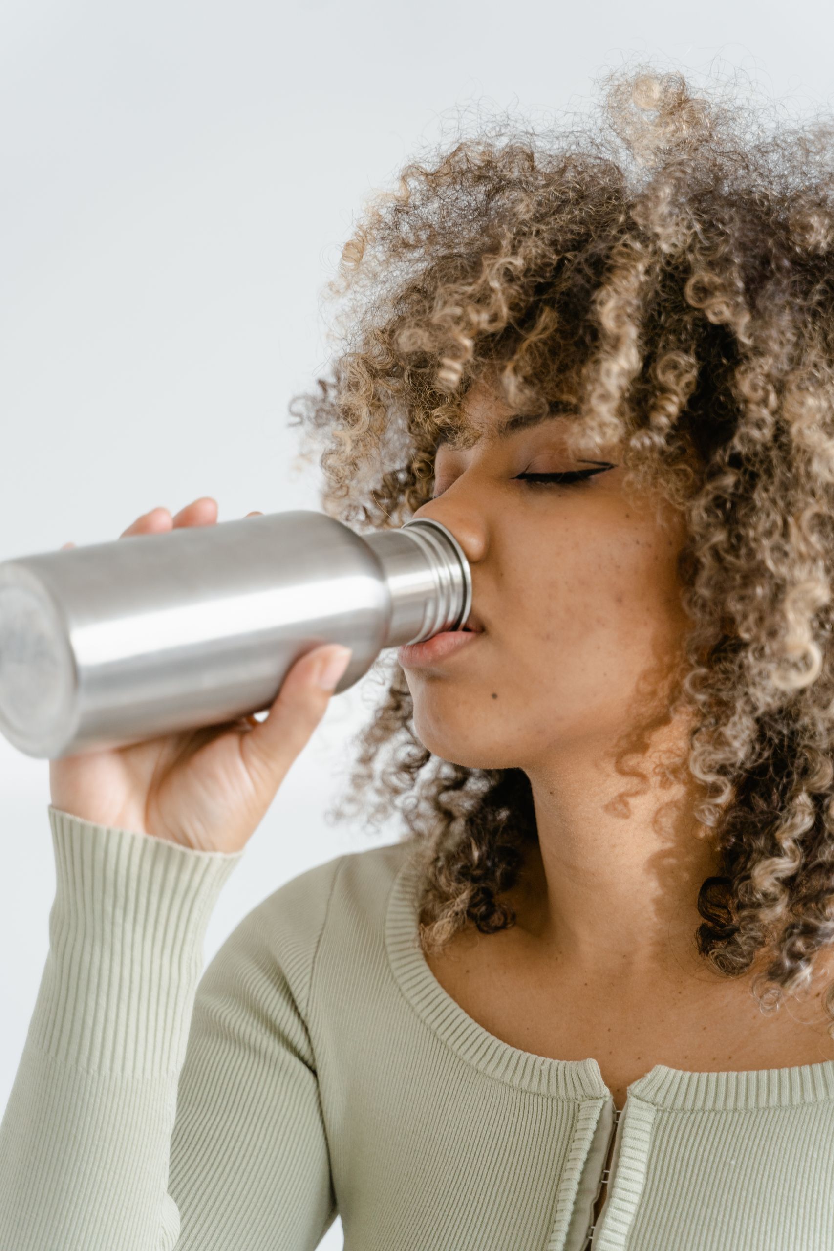 Woman with curly hair drinking from a stainless steel bottle in a close-up view.