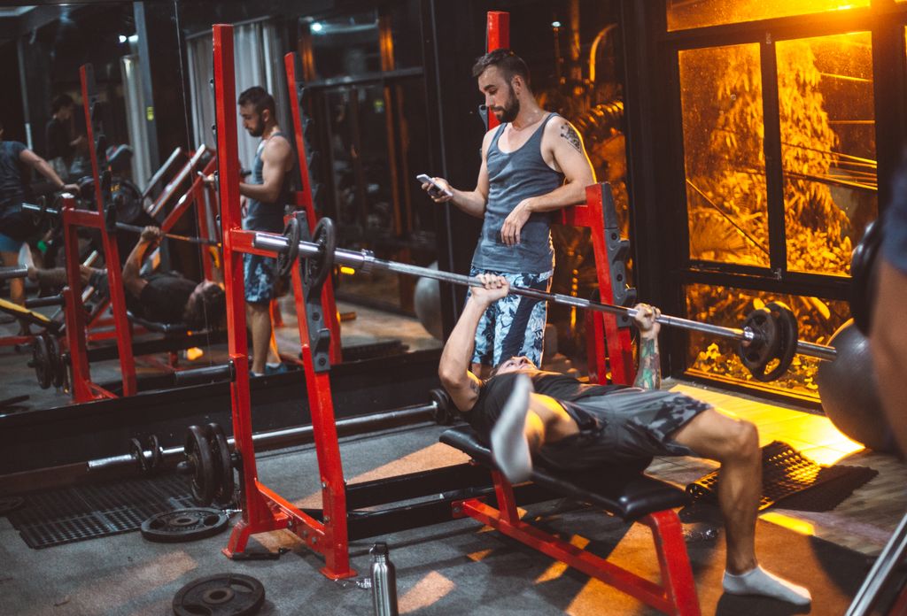 Full body concentrated sporty fitness trainer using smartphone while male on bench struggling with heavy barbell in fitness club gadget addiction concept