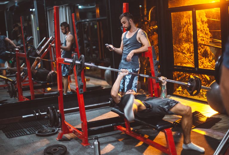 Full body concentrated sporty fitness trainer using smartphone while male on bench struggling with heavy barbell in fitness club gadget addiction concept