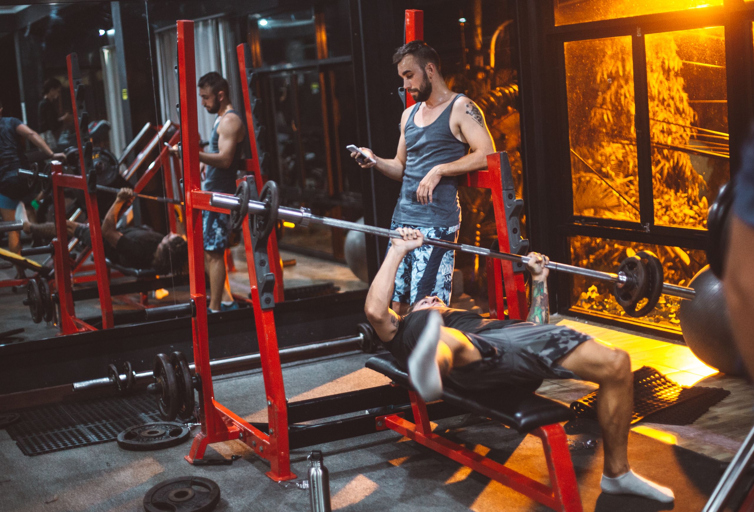Full body concentrated sporty fitness trainer using smartphone while male on bench struggling with heavy barbell in fitness club gadget addiction concept