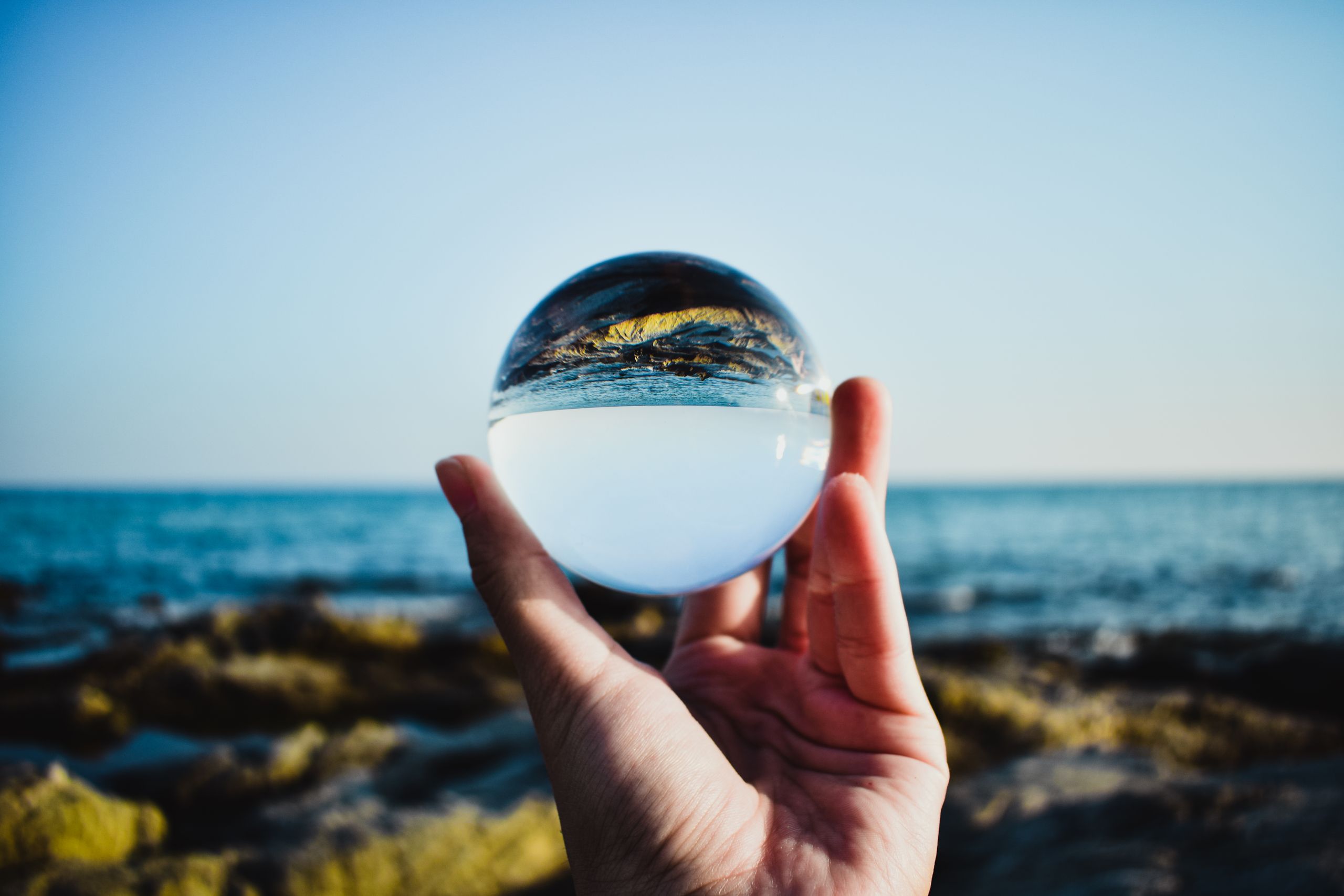A hand holds a glass ball reflecting the beach and ocean horizon.